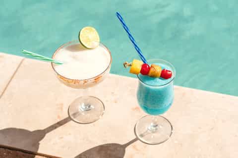 Two colorful cocktails by turquoise pool with tropical garnishes and fruit skewers