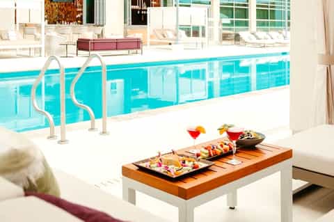 Modern indoor pool with glass enclosure, white lounge chairs, tropical plants, and appetizers on wooden table