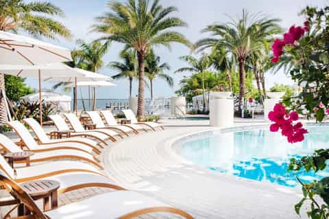 Luxury resort pool area with palm trees, white umbrellas, lounge chairs, and ocean view in background