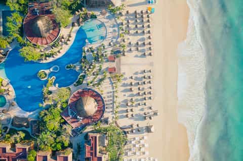 Aerial view of beachfront resort with pools, umbrellas, palm trees, and turquoise ocean