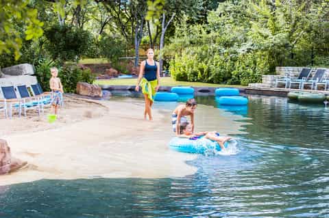 Family enjoying natural spring water with float tubes, sandy beach, trees, and resort facilities