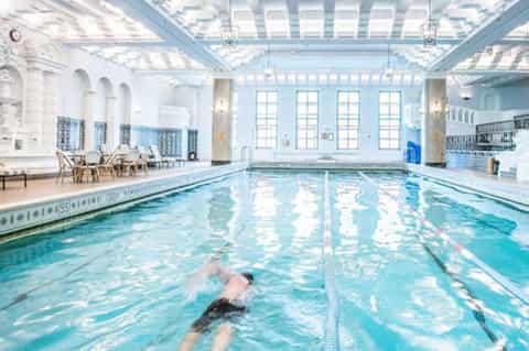 Indoor Olympic-length lap pool with skylight ceiling, lane markings, and swimmer doing freestyle