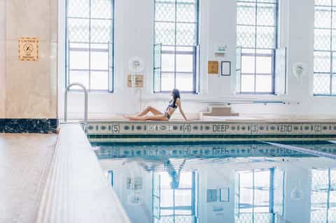 Indoor lap pool with depth markers and lane dividers, woman sitting on pool edge with windows behind