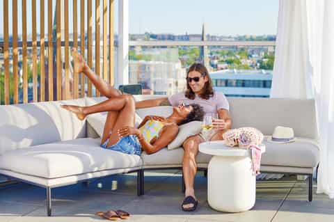 Couple relaxing on modern rooftop lounge with city skyline view, white furniture, and afternoon sun