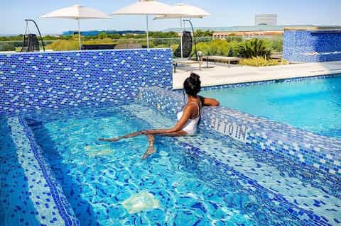 Woman relaxing in bright blue tiled resort pool under white umbrellas with modern architecture