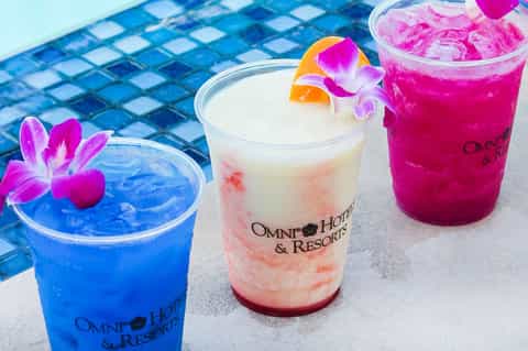 Three colorful tropical cocktails by a pool with vibrant flowers