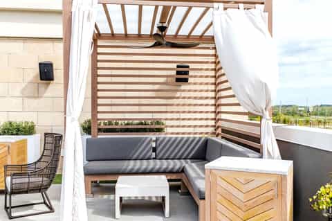 Modern outdoor living space with gray sectional sofa under wooden pergola with white curtains and ceiling fan