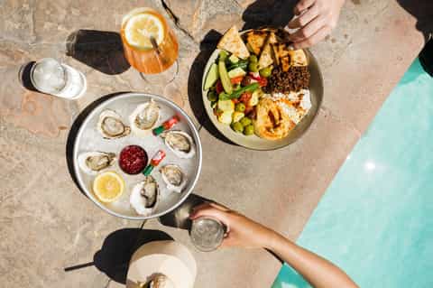 Overhead view of oysters, ceviche bowl, and cocktails on poolside concrete deck