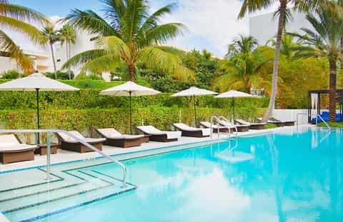Contemporary pool area with white lounge chairs, cream umbrellas, palm trees, and white residential buildings