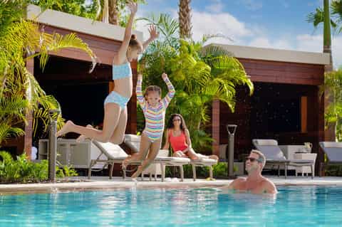 Family enjoying resort pool with children splashing, palm trees, and modern cabana structures