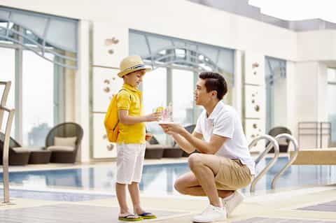 Adult and child in yellow clothing by indoor pool with floor-to-ceiling windows overlooking city skyline