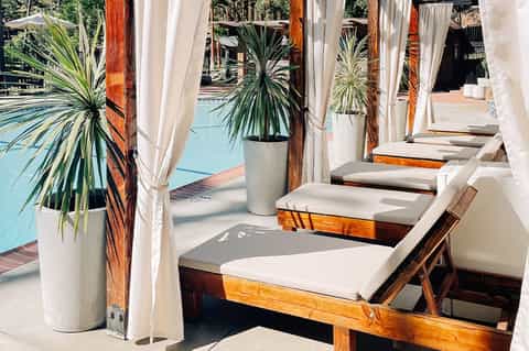Poolside cabanas with white curtains, palm plants, and lounge chairs overlooking a turquoise swimming pool