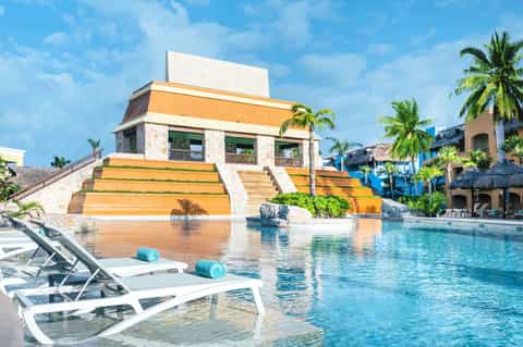 Resort pool with yellow pyramid structure, white loungers, palm trees, and turquoise water