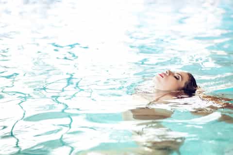 Woman floating peacefully in clear turquoise swimming pool water with natural sunlight reflections