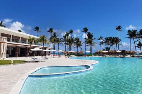 Luxury beachfront resort pool with modern building, white umbrellas, lounge chairs, and palm trees