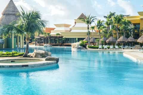 Tropical resort with large lagoon pool, thatched roof structures, palm trees, and modern yellow building