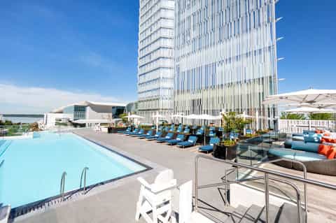 Modern hotel pool deck with blue loungers, white umbrellas, and high-rise building overlooking water