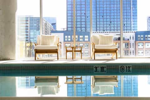 Modern lounge chairs by indoor pool overlooking city skyline and tall buildings