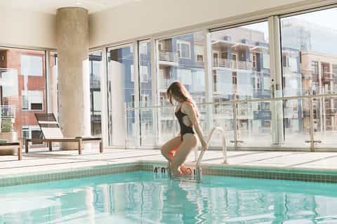 Woman about to enter indoor pool with large windows overlooking residential buildings
