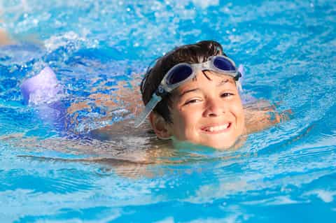 Young boy swimming in clear blue pool wearing goggles and smiling