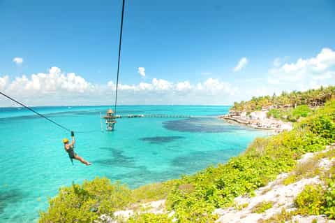 Person zip-lining over turquoise ocean water with palm trees and beach resort visible below