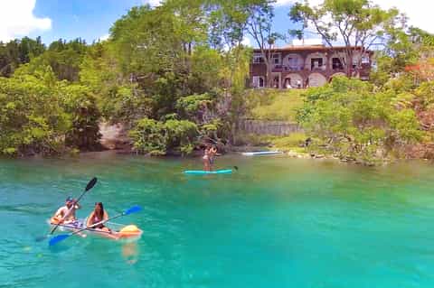 People paddleboarding in turquoise river near red brick building surrounded by lush green vegetation