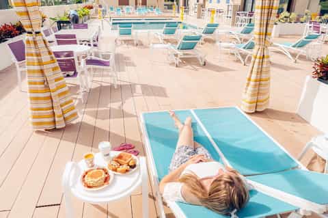 Woman relaxing on lounge chair with food tray overlooking pool deck