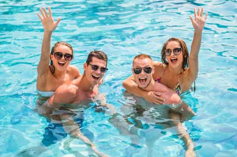 Four young adults enjoying themselves in a bright blue swimming pool, raising their arms and smiling