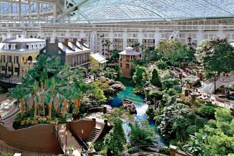 Indoor tropical garden atrium with glass ceiling, lush vegetation, water features, and colonial-style buildings