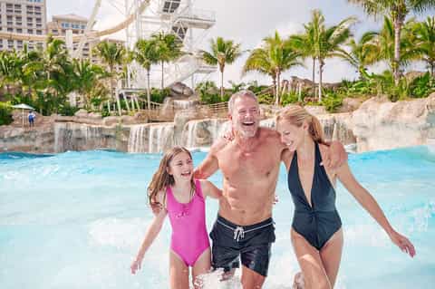 Family enjoying turquoise lagoon pool with waterfall feature and resort buildings with palm trees in background