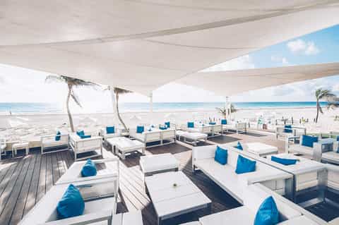 Modern beach club with white loungers under shade sails overlooking sandy beach and ocean