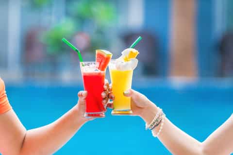 Two colorful cocktails with straws held up against blue pool background