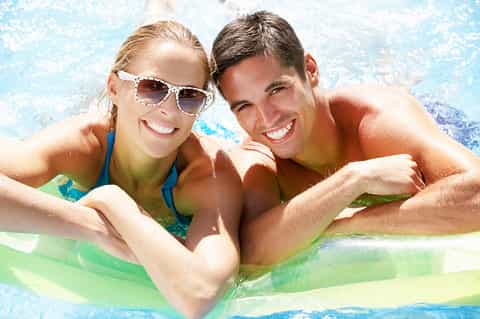Young couple in pool float smiling at camera in bright blue swimming pool