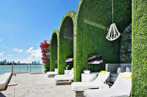 Upscale waterfront lounge with green ivy-covered structures, white loungers, and city skyline view