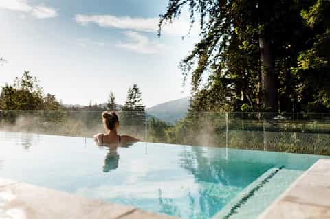 Woman relaxing in steaming infinity pool with mountain forest views and glass railing