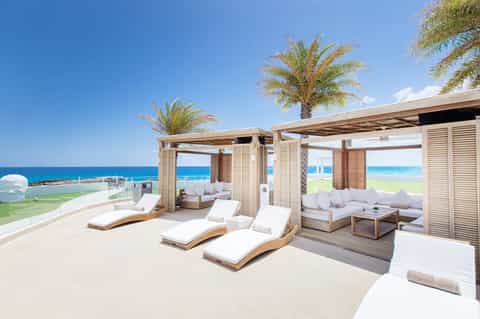Oceanfront terrace with wooden loungers under pergolas, palm trees, and blue water beyond
