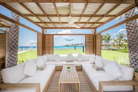 Beachfront cabana with white cushioned seating, wooden pergola, ocean views, and palm trees