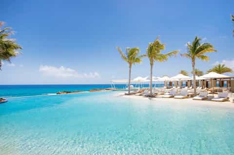 Tropical beach resort with turquoise water, palm trees, white lounge chairs, and beige umbrellas