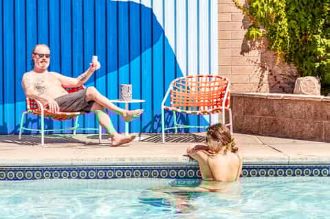 Man relaxing poolside in chair while woman swims in bright turquoise pool with blue wall mural