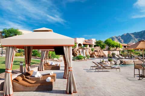 Desert resort pool area with cabanas, lounge chairs under umbrellas, and mountains in background