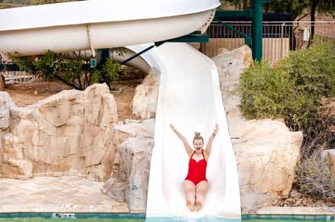 Child on white water slide at resort, splashing into pool with rocky landscape and sailboat visible.