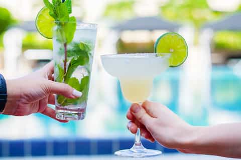 Two hands holding refreshing mojito and margarita cocktails by a swimming pool
