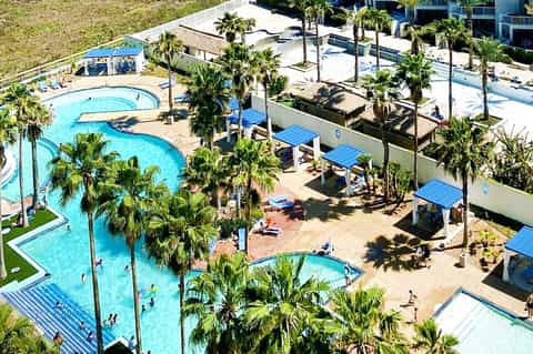 Aerial view of large resort pool complex with palm trees, blue umbrellas, and multiple pools