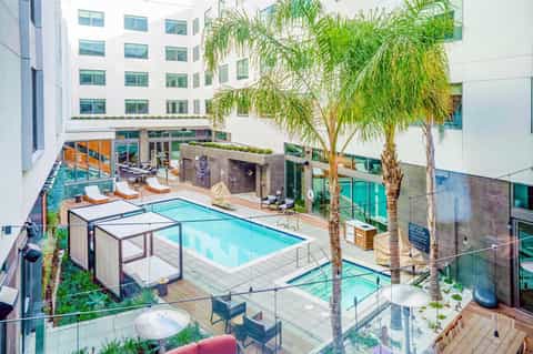 Resort courtyard with swimming pool, lounge chairs, palm trees, and modern white building
