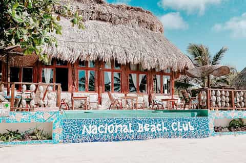 Colorful beachfront beach club with red wooden structure, thatched roof, and turquoise mosaic tile signage