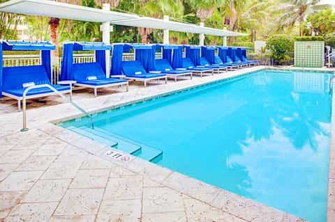 Resort swimming pool with crystal-clear water, blue cabanas with curtains, and palm tree landscaping