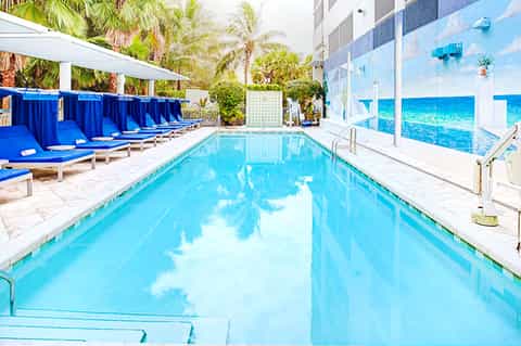 Indoor lap pool with blue water, palm trees, blue cabanas, and large windows overlooking waterfront