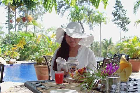 Woman reading poolside in tropical setting with fresh beverages, flowers, and lush garden landscape
