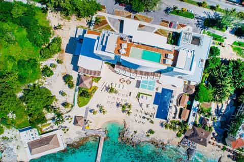 Aerial view of beachfront resort with white and blue buildings, pools, beach, and lush vegetation