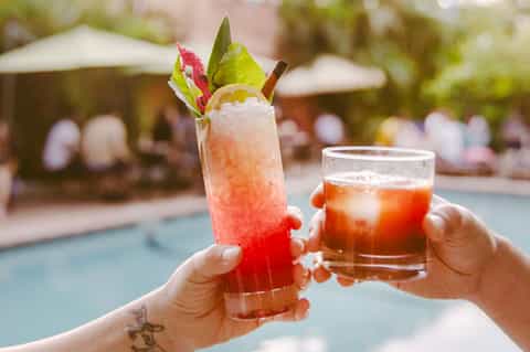 Two refreshing cocktails with fruit garnishes held by pool with umbrellas